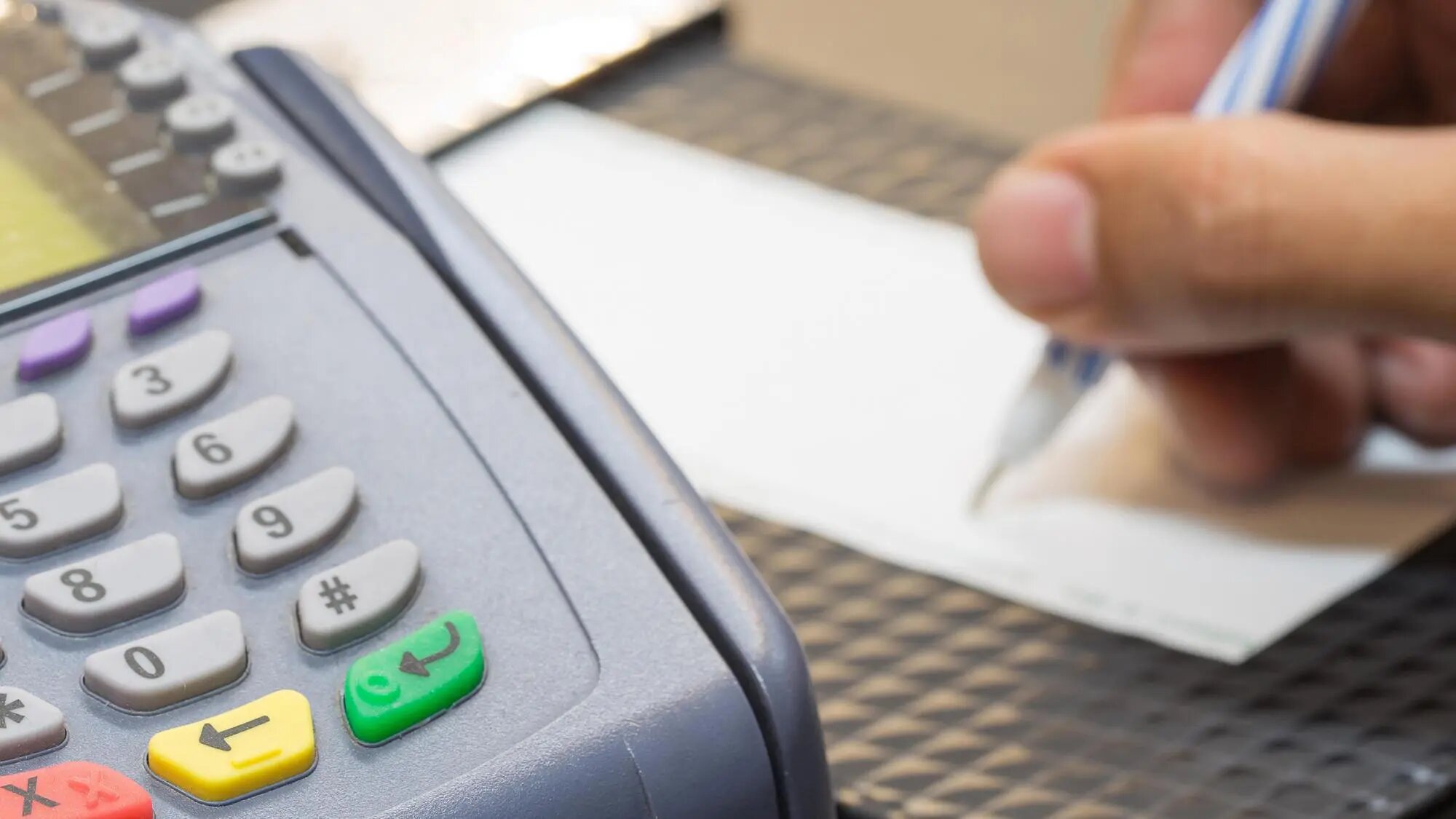 person writing their signature on a receipt next to a credit card reader